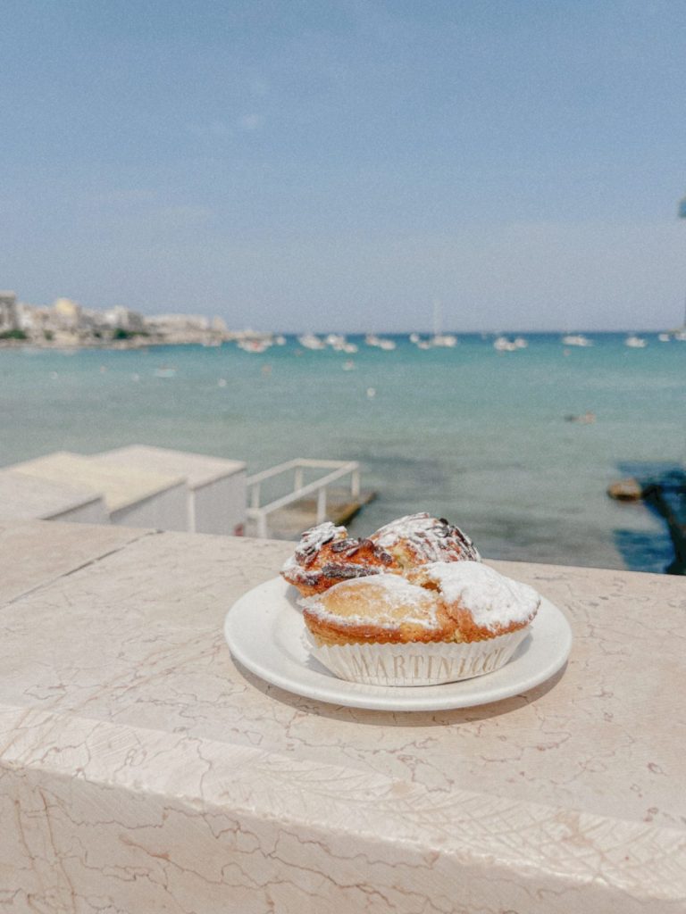 Pasticciotti spolverati di zucchero a velo su un piatto, con il mare del Salento sullo sfondo